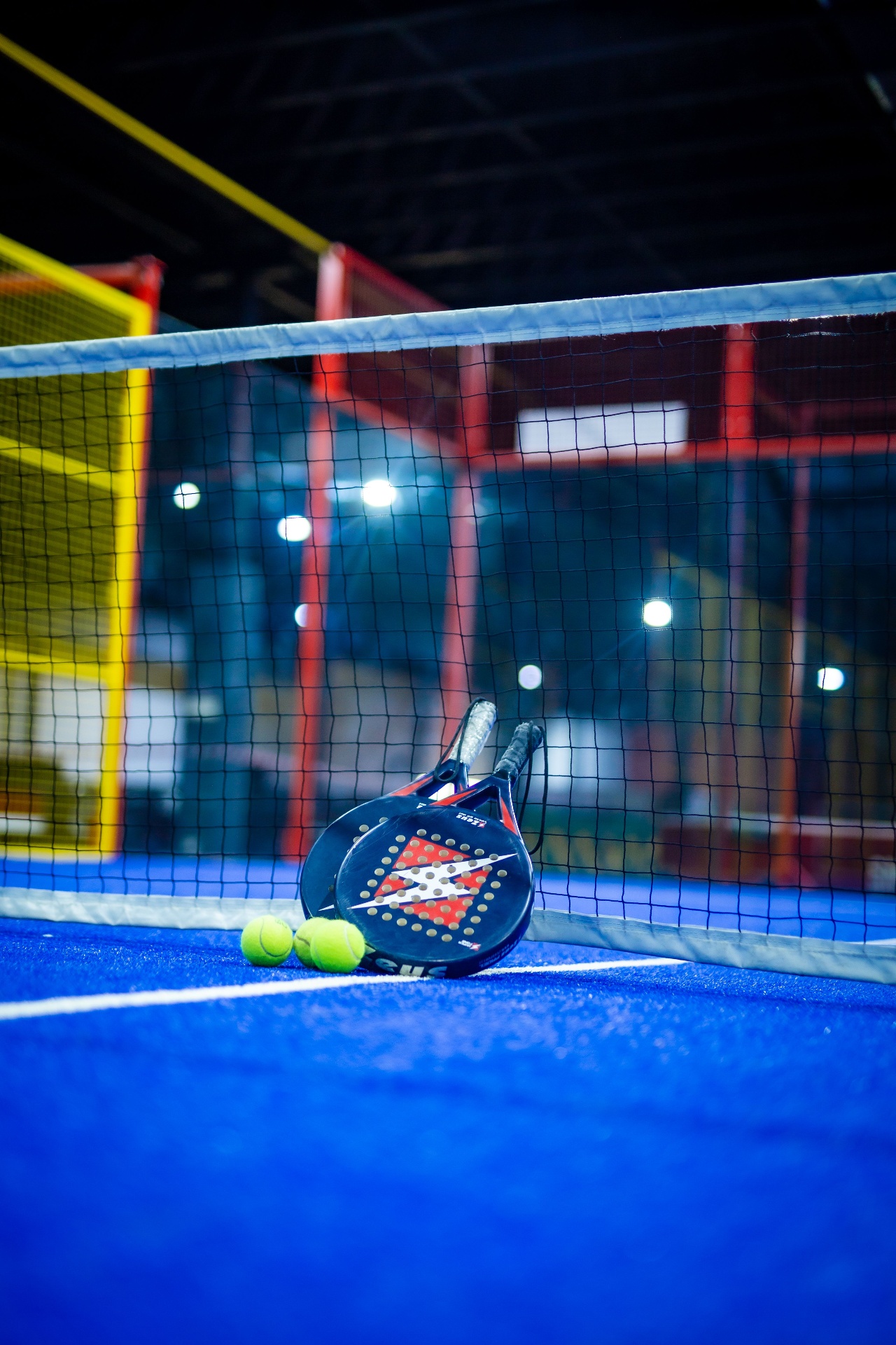 Two padel rackets and balls leaning against the net on an indoor padel court
